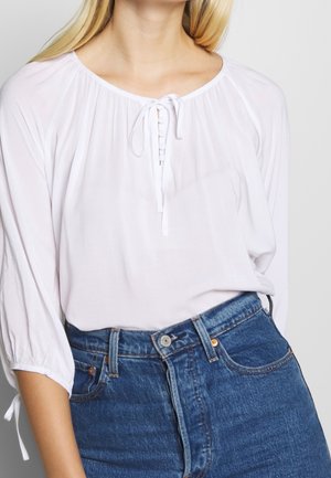 Mujer con una blusa blanca de manga larga, holgada, con botones al frente y lazo, metida dentro de unos jeans de mezclilla azul de cintura alta.