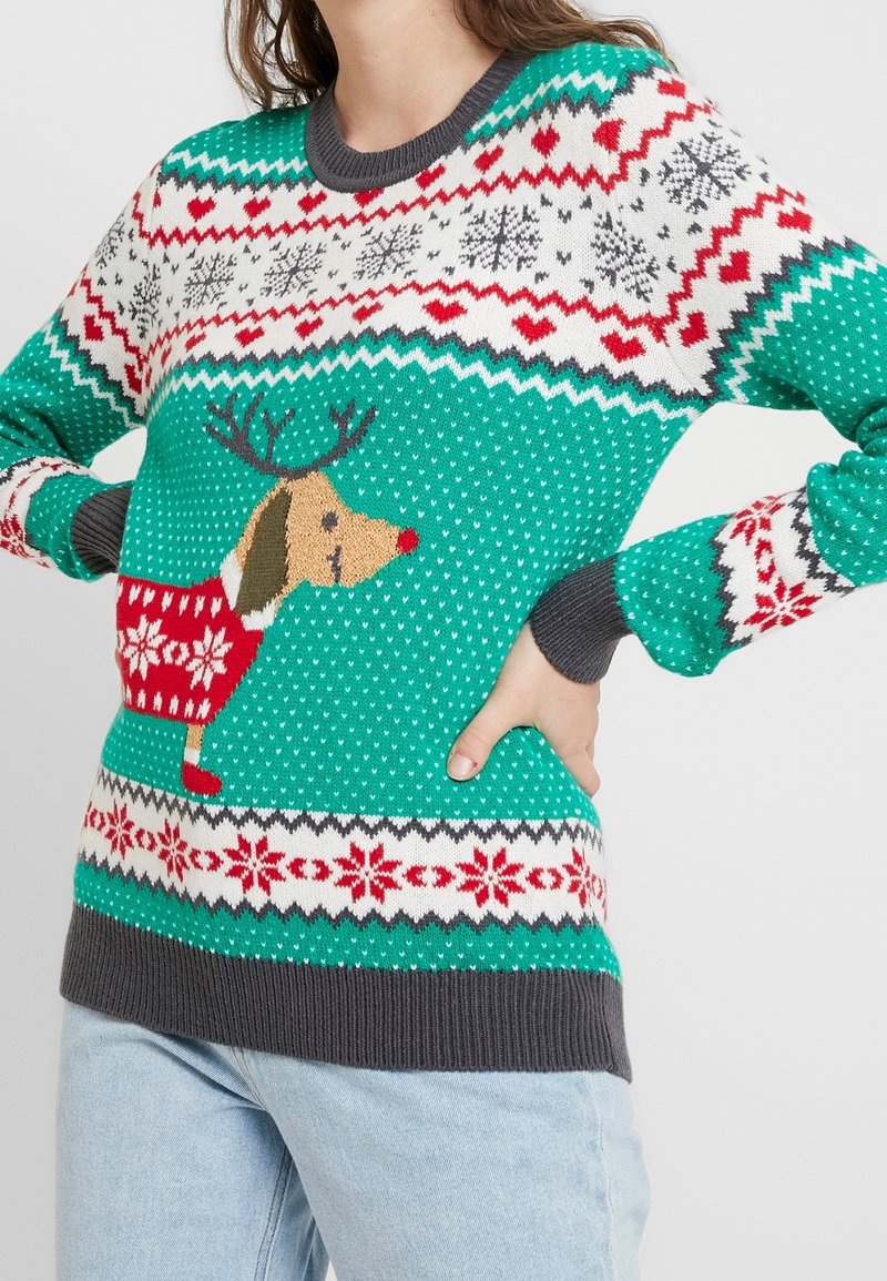 Femme portant un pull de Noël turquoise avec des motifs de rennes, de cœur rouge, de flocons de neige et des poignets gris, associé à un jean bleu clair.