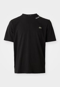 Svart T-skjorte med korte ermer og rund hals, med en liten grønn krokodille-logo på brystet og hvit kursiv tekst "Lacoste" nær kragen.
