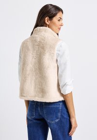 Gilet in finta pelliccia color beige chiaro con una finitura morbida e texture. Presenta un colletto alto e orlo dritto, abbinato a jeans in denim blu.