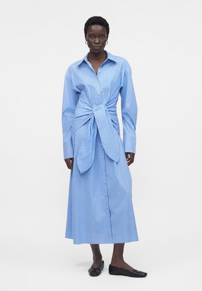 Γυναίκα που φοράει μακρύ μπλε ριγέ φόρεμα με κουμπιά, με φαρδιές μανσέτες και έναν μεγάλο δεμένο κόμπο στη μέση, συνοδευόμενο από μαύρα φλατ παπούτσια.