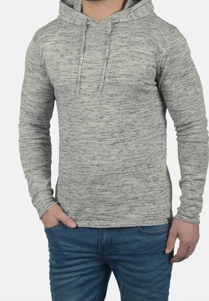 Man wearing light gray speckled long-sleeve hoodie with drawstrings and blue jeans, standing with one hand in pocket.