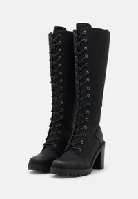 Bottes noires montantes jusqu'au genou en matériau lisse, avec des lacets à l'avant, des œillets en métal, un talon épais et une semelle texturée pour l'adhérence.