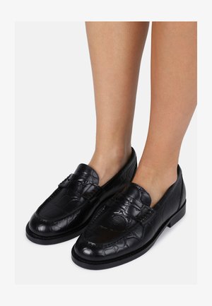 Mocassini neri in pelle con texture a coccodrillo, con punta arrotondata e senza lacci, indossati su gambe nude. Suola piatta e tacco basso.