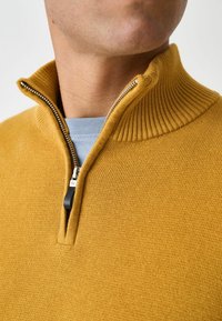 Gelber Pullover mit Reißverschluss am Hals, ausgestattet mit einem halben Reißverschluss und einem strukturierten Strickmuster. Leicht und lässig geschnitten, über einem blauen Hemd getragen.