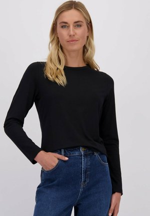 Blonde vrouw in een zwart lange mouw shirt, ingestopt in een hoge taille blauwe jeans, staand met één hand in haar zak tegen een effen achtergrond.