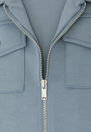 Lyseblå lynlåstrøje med sølvfarvet lynlås ned gennem midten, to brystlommer og et tekstureret stof.