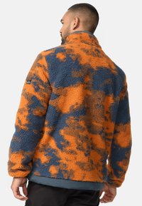 Fleece dzseki narancs és kék álcázó mintával, texturált anyaggal, magas gallérral és egy címkével a bal ujjon.