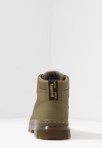 Bota alta marrón con suela translúcida gruesa, costura amarilla y tirador negro que dice "CON SUELAS ELÁSTICAS", vista desde la parte de atrás.