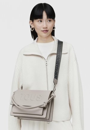 Bolso de hombro de cuero gris con logo "TOUS" en relieve, asa anudada y una correa texturizada con branding repetido.