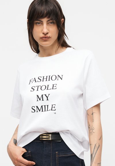 Νεαρό άτομο με σκουρόχρωμα μαλλιά φοράει λευκό μπλουζάκι με το κείμενο «FASHION STOLE MY SMILE», σκούρο τζιν, μαύρη ζώνη και δείχνει πολλά τατουάζ στα χέρια.