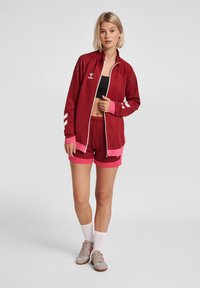 Chaqueta deportiva roja con cierre blanco y acentos rosas, combinada con pantalones cortos rojos. La tela muestra textura; incluye logotipo y detalles a rayas.