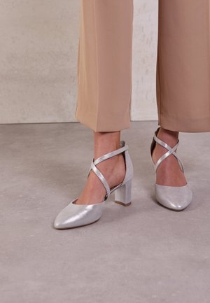 Srebrne szpilki z wąskim noskiem, o fakturowanym wykończeniu, z krzyżującymi się paskami na kostkach, w połączeniu z beżowymi spodniami w kant.