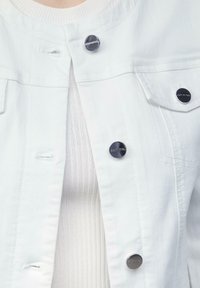 Veste en denim blanc avec des boutons-pression en métal foncé, comportant deux poches poitrine et une texture légèrement usée. Superposée sur un haut à côtes clair.