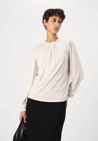 Lys beige langermet topp med rynkede detaljer foran og flytende ermer, båret over et svart skjørt, laget av myk, glatt stoff.