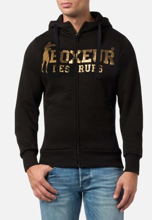 Felpa nera con zip, caratterizzata da un logo frontale in oro, cappuccio con cordino e tasche a marsupio. Il tessuto risulta morbido con una texture liscia.