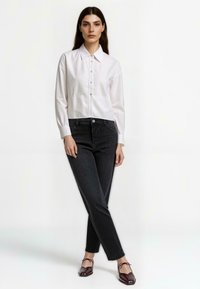 Biała koszula z kołnierzykiem z guzikami, połączona z ciemnymi dżinsami o wysokim stanie. Modelka ma na sobie głębokie burgundowe płaskie buty, prezentując elegancki, nowoczesny wygląd.