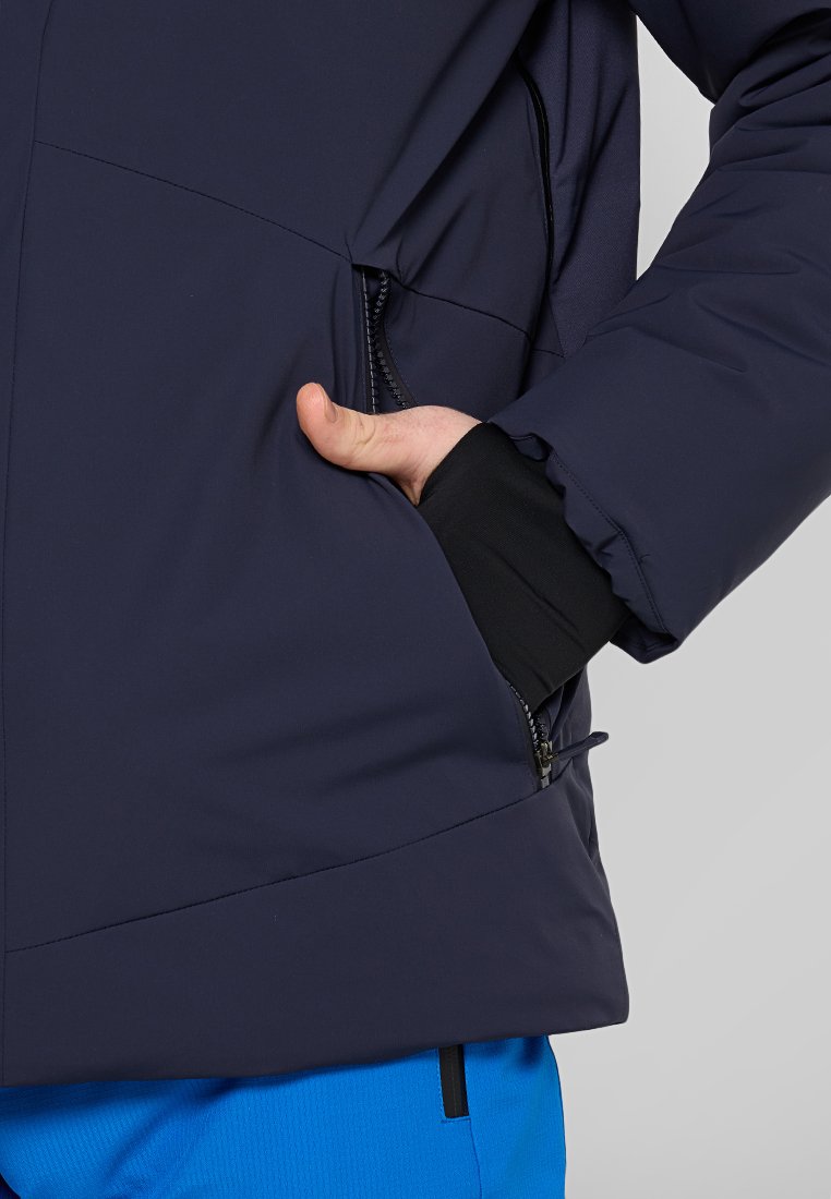 Giacca isolata blu navy con tasca con zip, caratterizzata da una texture liscia e un design aderente, abbinata a pantaloni blu brillante.