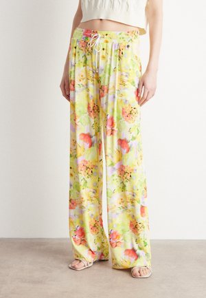 Kvinde iført løse bukser med vide ben og et mønster af gule, pink og grønne blomster og frugter, kombineret med en cremefarvet strikket cropped top og hvide sandaler.