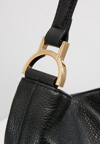 Mustamassa nahkalaukussa on kultasävyisiä yksityiskohtia ja teksturoitu pinta, ja se on suljettu erottuvalla pyöreällä koukulla.