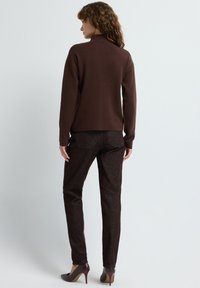 Vrouw staat, met de rug naar de camera, gekleed in een bruine coltrui, donkerbruine broek en donkerbruine pumps op een effen lichte achtergrond.