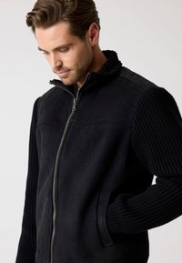 Schwarze Jacke zum Reißverschluss mit Fleecematerial am Körper, gerippten Strickärmeln und hohem Kragen. Verfügt über Seitentaschen und eine glatte Textur.