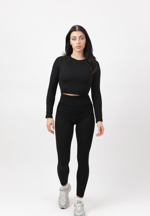 Donna che cammina in avanti indossando un top corto a maniche lunghe nero, leggings a vita alta e sneakers bianche su sfondo bianco uniforme.