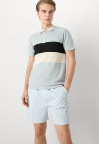 Polo de manga curta em azul claro, com riscas horizontais em preto, creme e azul escuro; fecho com zip e colarinho canelado; acompanhado de calções à risca.
