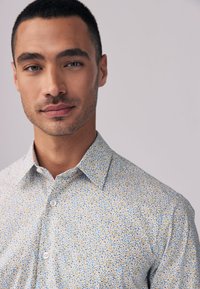 Lyseblå skjorte med knapper og blomsterprint samt en spredt krave, med et tekstureret mønster i gule og blå accenter. Blødt stoffinish.