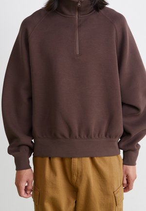 Person trägt einen braunen Quarter-Zip-Sweatshirt mit elastischem Saum und beige Cargohosen, steht vor einem schlichten hellen Hintergrund.