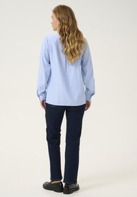 Camicia a righe blu chiaro con bottoni e maniche lunghe, abbinata a jeans blu scuro e mocassini neri. Texture del tessuto liscia visibile.
