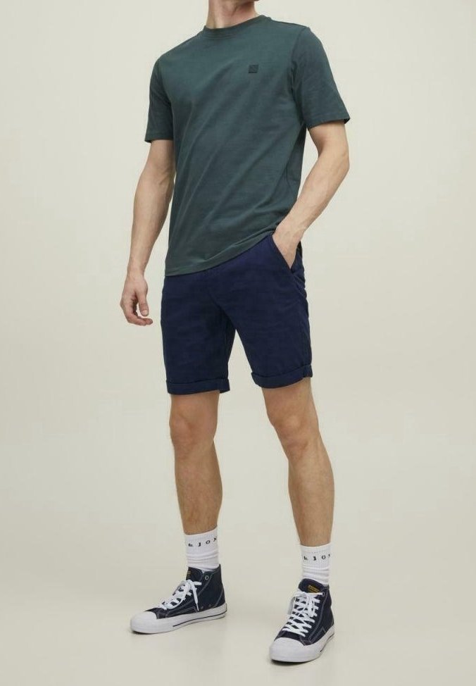 Homme portant un t-shirt à manches courtes vert foncé, un short bleu marine retroussé, des chaussettes blanches montantes et des baskets montantes bleu marine, debout avec une main dans la poche.