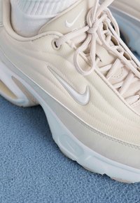 Nike sportiniai bateliai švelniai bežinės spalvos, su tekstūruota medžiaga, lygiais akcentais ir ryškiu logotipu. Šviesios spalvos padas su matomais amortizacijos detalėmis.