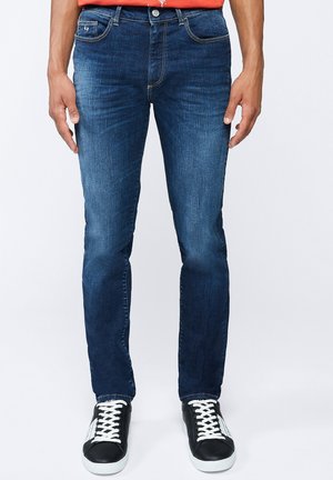 Jeans aderenti in denim blu con leggero effetto scolorito, cuciture arancioni a contrasto, cinque tasche e una cintura scura. Abbinati a sneakers nere.