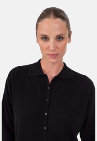 Cardigan noir boutonné avec col, fabriqué en tissu doux. Le design présente une coupe décontractée et une texture subtile, idéal pour superposer.