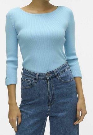 Kvinde iført en lyseblå ribstrikket top med trekvartlange ærmer, stukket ned i højtaljede blå jeans mod en ensfarvet baggrund.