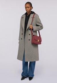 Grå dubbelknäppt ullrock med stora svarta knappar, tillsammans med en svart hoodie, utsvängda blå jeans och en bourgogny ransel.