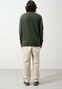 Dunkelgrüner Pullover mit hohem Kragen, lockerer Passform und Rippbund, kombiniert mit cremefarbigen, weit geschnittenen Hosen und dunklen Schuhen.