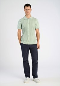 Hellgrüner, gestrickter Cardigan mit kurzen Ärmeln, strukturiertem Muster, Knopfverschluss und Rippbündchen, kombiniert mit dunkeln Marinehosen und weißen Turnschuhen.