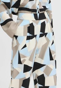 Pantalones con un patrón geométrico en tonos de azul claro, beige y negro, que presentan una tela texturizada, pliegues y un cinturón a juego en la cintura.
