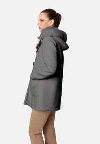 Abrigo de lana gris con capucha, con un forro de pelaje sintético, bolsillos laterales y un corte holgado, combinado con pantalones de color marrón claro.
