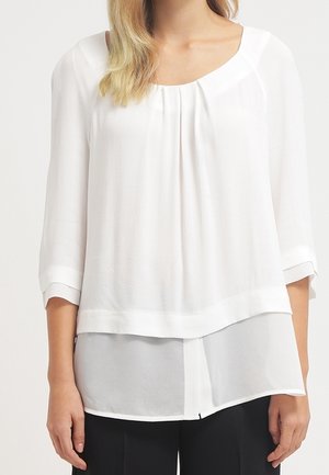 Blusa bianca semi-trasparente con scollo rotondo, maniche lunghe e design a strati. Presenta sottili pieghe e un orlo che si trova al di sotto della linea della vita.