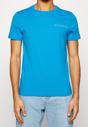 Mężczyzna ubrany w jasnoniebieską koszulkę z krótkim rękawem z napisem "HILFIGER" na piersi oraz jasnoniebieskie dżinsy na jednolitym tle.