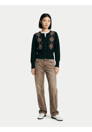 Femme debout portant un cardigan noir boutonné avec un motif losanges beige, un jean marron à jambes droites et des chaussures blanches sur un fond uni.