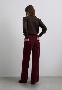 Pantalones anchos de pana color granate con bolsillos traseros, combinados con un suéter marrón oscuro con mangas abullonadas, vistos desde atrás.