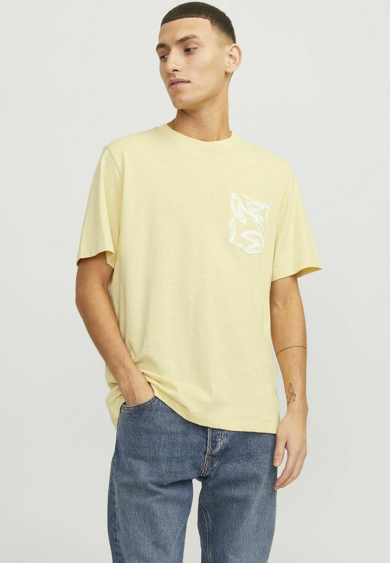 Lys gul kortærmet T-shirt med en frontlomme med et hvidt mønsterdesign. Stof har en blød tekstur, kombineret med blå denimjeans.