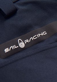 Laivastonsininen kangas, jossa on sileä pinta, ja musta etiketti, jossa valkoiset tekstit "SAIL RACING" sekä pyöreä logo.