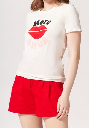 Frau trägt ein weißes T-Shirt mit roten Lippen und dem Schriftzug „More Amor“, kombiniert mit hellroten plissierten Shorts.