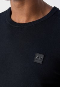 Gros plan d'une personne portant un pull col rond bleu marine foncé avec un petit patch logo carré noir sur la poitrine.