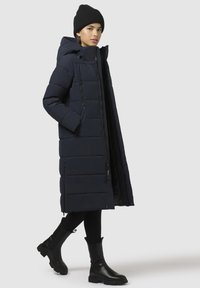 Cappotto lungo puffer blu navy con cappuccio, tasche con zip e motivo trapuntato. Abbinato a stivali neri fino al ginocchio e leggings.